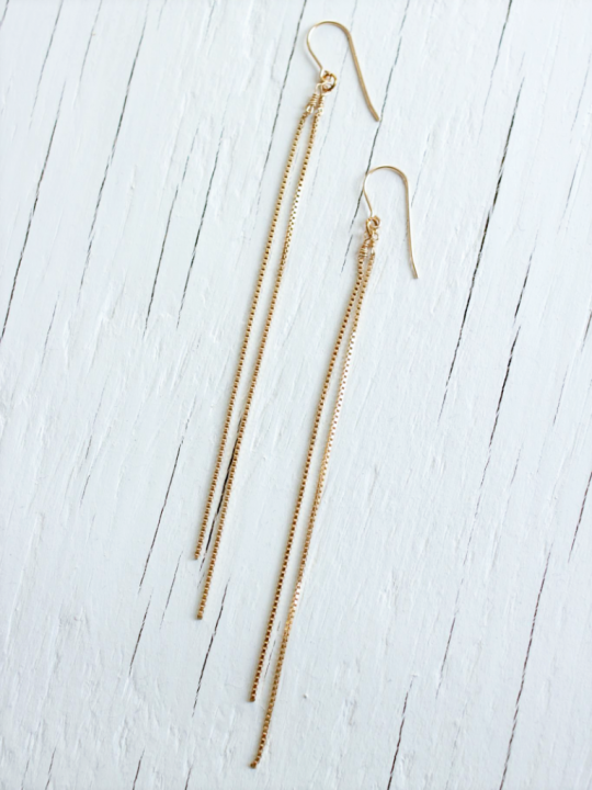 Two earrings made with 2 long gold chains each strung from a gold earwire sit on top of a white wood background