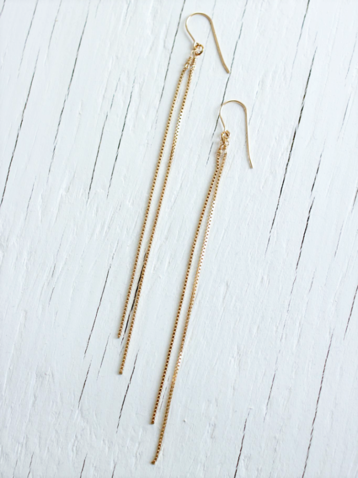 Two earrings made with 2 long gold chains each strung from a gold earwire sit on top of a white wood background