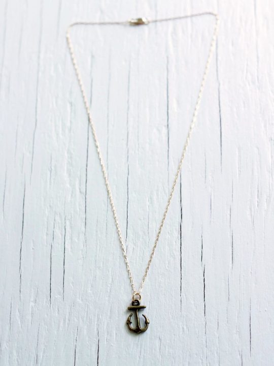Nautical necklace featuring a brass anchor on a 14kt gold filled chain laying on top of a white wooden background