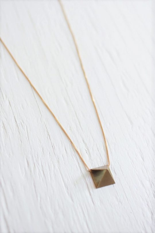 Gold pyramid necklace with raw brass pyramid pendant strung on 14kt gold fill chain laying on white wooden background