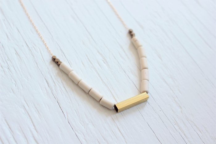 Beaded necklace with white tube beads and a brass tube on a gold chain, laying on a white wood background