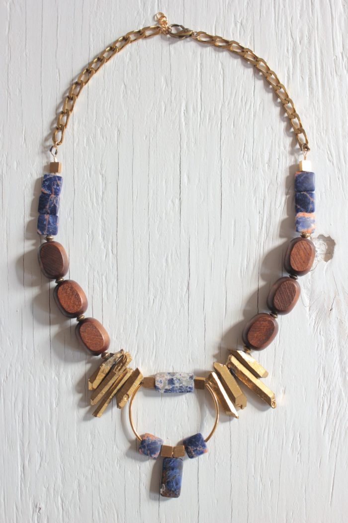 Bib necklace features blue sodalite stones, wooden beads, gold dipped prisms and geometric brass components