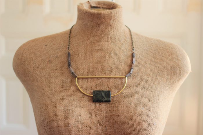 Modern necklace with a green jasper focal stone, curved brass bars, rectangle stones on a brass chain on a mannequin in front of a beige wall